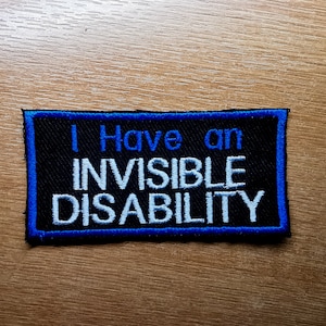 Puede incluir: Un parche negro con borde azul que dice "Tengo una discapacidad invisible" en letras blancas.