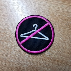 Peut inclure: Un écusson brodé noir et rose avec un cintre blanc barré d'une ligne rose. L'écusson est un symbole d'interdiction de cintre.