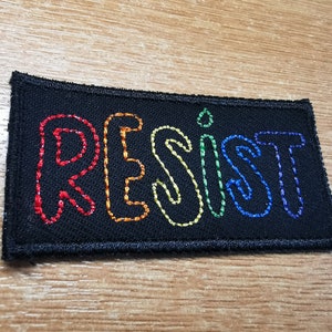 May include: A black fabric patch with the word "RESIST" embroidered in rainbow colors. The letters are outlined in white.