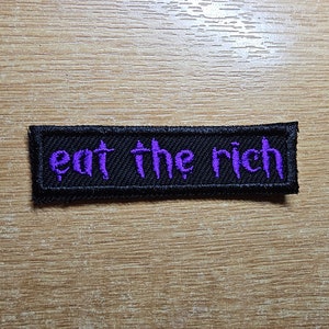 May include: Black fabric patch with the words "eat the rich" embroidered in purple.