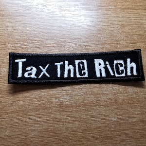 May include: Black fabric patch with white embroidered text that reads "Tax The Rich".