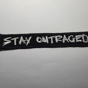 May include: Black rectangular embroidered patch with the words "STAY OUTRAGED" in white, stylized lettering. The patch has a black border and is likely designed to be sewn or ironed onto clothing or accessories.