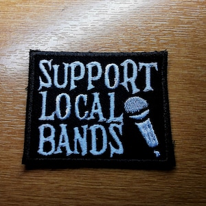 Peut inclure: Un écusson brodé noir avec du texte blanc qui dit "Support Local Bands" et un graphique de microphone blanc.