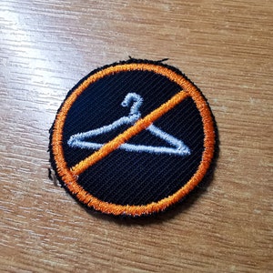 Puede incluir: Un parche bordado negro y naranja con una percha blanca tachada. El parche es una señal de prohibido colgar.