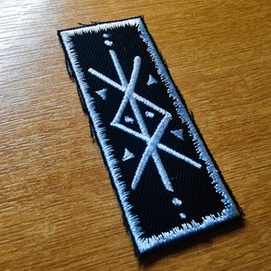 May include: White embroidered symbol on a black patch. The symbol is a stylized design of intersecting lines and triangles.