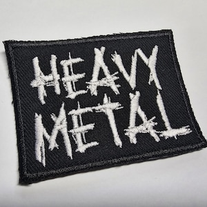 May include: Black embroidered patch with the words "HEAVY METAL" in white, stylized lettering. The patch is rectangular with a black border and is likely designed to be sewn onto clothing or accessories.