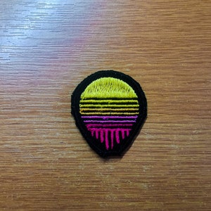 Puede incluir: Un parche bordado negro y amarillo con un diseño de puesta de sol. La puesta de sol es amarilla, naranja y rosa con un efecto de goteo.