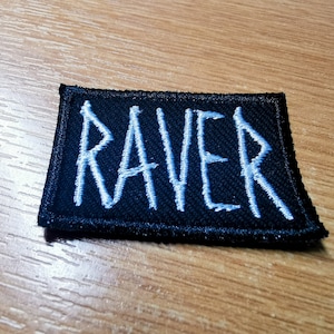 Op de afbeelding: Zwart rechthoekig embleem met het woord "RAVER" geborduurd in lichtblauwe draad. De patch heeft een zwarte rand en ligt op een houten oppervlak.