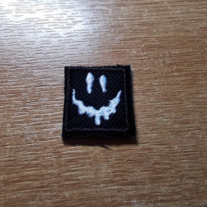 May include: A black fabric patch with a white embroidered smiley face. The smiley face has a dripping effect.
