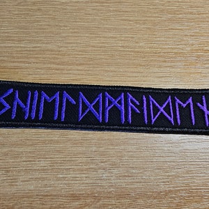May include: A rectangular black patch with purple embroidered runes. The patch is on a wooden surface. The runes spell out a word or phrase. The patch is likely for clothing or accessories.