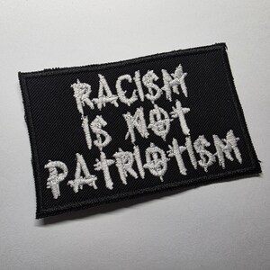 May include: A black rectangular patch with white embroidered text that reads "RACISM IS NOT PATRIOTISM". The patch has a stitched border and is likely designed to be sewn onto clothing or accessories.