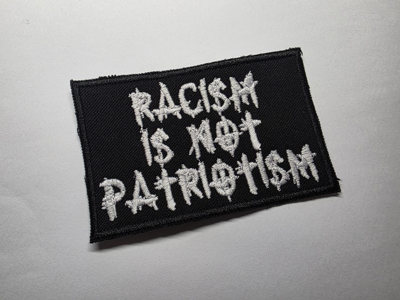 May include: A black rectangular patch with white embroidered text that reads "RACISM IS NOT PATRIOTISM". The patch has a stitched border and is likely designed to be sewn onto clothing or accessories.