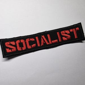 May include: A black rectangular patch featuring the word "SOCIALIST" in red, stencil-style lettering. The patch has a black border and is set against a white background. The text is centrally aligned on the patch.