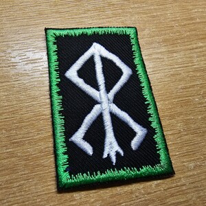 May include: A white embroidered symbol on a black patch with a green border. The symbol is a stylized rune.