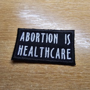 Op de afbeelding: Zwart rechthoekig embleem met witte geborduurde tekst: "ABORTION IS HEALTHCARE". Het embleem heeft een zwarte rand en staat op een houten achtergrond. Het embleem is waarschijnlijk voor kleding of accessoires.