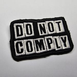 Könnte beinhalten: Schwarzer rechteckiger Aufnäher mit weiß gesticktem Text „DO NOT COMPLY“. Der Aufnäher hat einen schwarzen Rand und ist wahrscheinlich zum Aufnähen oder Aufbügeln auf Kleidung oder Accessoires bestimmt.