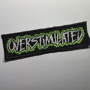 May include: Black rectangular patch with the word "OVERSTIMULATED" embroidered in white, outlined with green. The patch has a green, spiky border. The text is in a bold, stylized font.