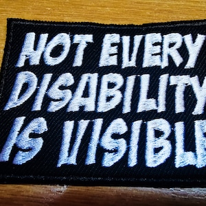 Könnte beinhalten: Schwarzer Stoff-Patch mit weiß gesticktem Text, der "Not every disability is visible" lautet.