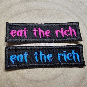 Puede incluir: Dos parches de tela negra con la frase "eat the rich" bordada en hilo rosa y azul.