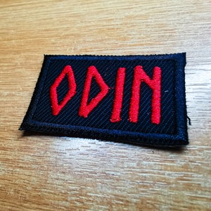 Puede incluir: Un parche de tela negro con letras bordadas en rojo que dicen "ODIN".