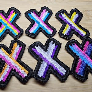 Puede incluir: Seis parches bordados para planchar en forma de 'X' con diferentes combinaciones de colores de blanco, rosa, azul, morado, amarillo y naranja.