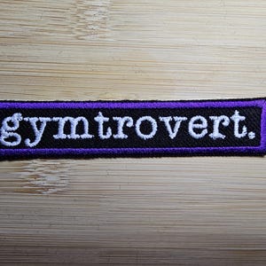 Op de afbeelding: Geborduurde patch met het woord "gymtrovert." in het wit, gestikt op een zwarte rechthoek met een paarse rand. De patch is rechthoekig en ontworpen om op kleding of tassen te strijken of te naaien.