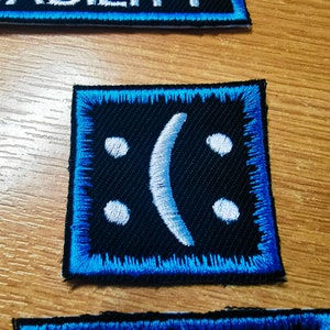 Può includere: Una toppa ricamata blu e nera con un viso triste bianco. La toppa ha un bordo blu ed è realizzata in tessuto strutturato.