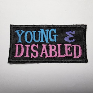 May include: A rectangular black embroidered patch featuring the text YOUNG in light blue, a purple ampersand symbol, and the word DISABLED in pink. The patch has a black stitched border and is designed for application to fabric items.