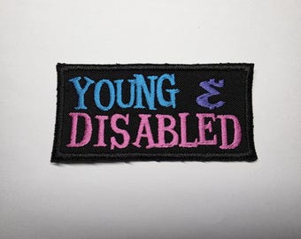 Op de afbeelding: Een rechthoekige zwarte geborduurde patch met de tekst YOUNG in lichtblauw, een paars ampersand-teken en het woord DISABLED in roze. De patch heeft een zwarte gestikte rand en is ontworpen voor bevestiging op stoffen artikelen.