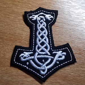 May include: A black embroidered patch with a white outline of a stylized hammer, a Norse symbol, with a braided design in the center.