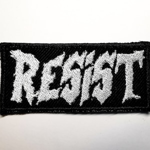 Puede incluir: Parche rectangular negro con la palabra "RESIST" bordada en blanco, con letras de estilo gótico. El parche tiene un borde negro y está diseñado para ser cosido en ropa o accesorios.