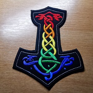 May include: A rainbow-colored embroidered patch depicting a stylized Mjolnir hammer with a Celtic knot design. The patch is black with white stitching around the edges.