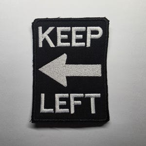 Op de afbeelding: Zwart rechthoekig embleem met wit geborduurde tekst en pijl. De tekst luidt "KEEP" boven een pijl die naar links wijst en "LEFT" eronder. Het embleem heeft een zwarte rand.
