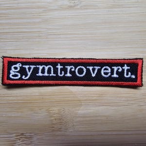 Op de afbeelding: Rode en zwarte geborduurde patch met de tekst "gymtrovert" in wit.