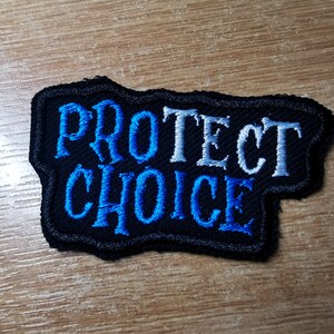 May include: Black embroidered patch with the words "PROTECT CHOICE" in white and blue thread.