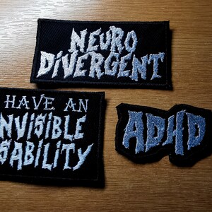 Pode incluir: Tr&ecirc;s patches de tecido preto com texto bordado branco. O patch superior diz "Neuro Divergente". O patch do meio diz "Eu tenho uma defici&ecirc;ncia invis&iacute;vel". O patch inferior diz "TDAH".