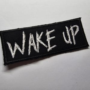 Può includere: Toppa rettangolare nera con le parole "WAKE UP" ricamate in bianco. Il testo è scritto in uno stile manoscritto. La toppa ha un bordo nero ed è progettata per essere cucita su vestiti o accessori.