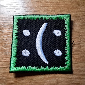 Può includere: Una toppa nera con bordo verde e un viso triste ricamato in bianco.