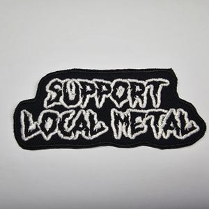 May include: A black embroidered patch featuring the text "SUPPORT LOCAL METAL" in white, bold lettering. The patch has a black border and is designed to be sewn or ironed onto clothing or accessories.
