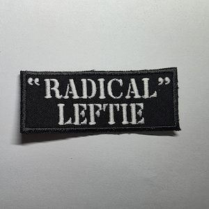 Könnte beinhalten: Schwarzer rechteckiger Aufnäher mit weiß gesticktem Text "RADICAL LEFTIE". Der Text ist in einer fetten, serifenlosen Schriftart gehalten und wird von Anführungszeichen umschlossen. Der Aufnäher ist wahrscheinlich aus Stoff und hat einen genähten Rand.