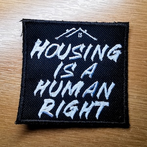 Puede incluir: Parche cuadrado negro con texto bordado blanco y un gráfico de una casa. El texto dice "HOUSING IS A HUMAN RIGHT". El parche está sobre una superficie de madera.