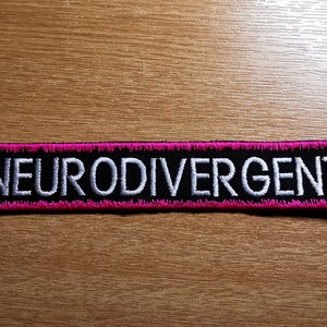 Op de afbeelding: Zwarte en roze geborduurde patch met de tekst "NEURODIVERGENT" in wit.