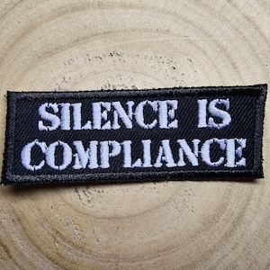 Könnte beinhalten: Schwarzer Stoff-Patch mit weiß gesticktem Text, der "Silence is Compliance" lautet.