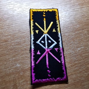 Puede incluir: Un parche negro bordado con un símbolo blanco y amarillo en el centro. El parche está bordeado de amarillo, blanco, rosa y morado.