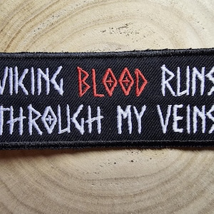 Op de afbeelding: Zwarte geborduurde patch met witte tekst die "Viking Blood Runs Through My Veins" luidt. Het woord "Blood" is rood.