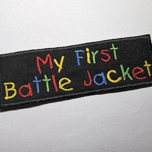 May include: Black rectangular patch embroidered with the words "My First Battle Jacket" in colorful thread. The text is in a playful, handwritten style, with each word in a different color: red, blue, green, and yellow.