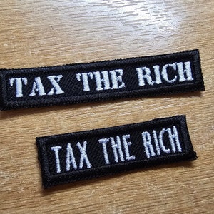 May include: Two black fabric patches with white embroidered text that reads "Tax the Rich". The patches are rectangular and have a slightly rough texture.