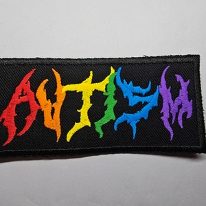 May include: A black fabric patch with the word "AUTISM" embroidered in rainbow colours. The letters are stylised in a jagged, metal-like font.