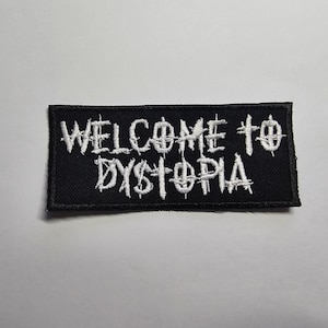 Könnte beinhalten: Schwarzer rechteckiger Aufnäher mit weiß gesticktem Text „WELCOME TO DYSTOPIA“. Der Text ist in einer verwitterten, gotischen Schriftart gehalten. Der Aufnäher ist für Kleidung oder Accessoires bestimmt.