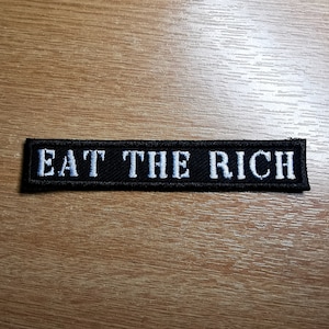 以下が含まれることがあります： 「EAT THE RICH」と白く刺繍された黒い布のパッチです。
